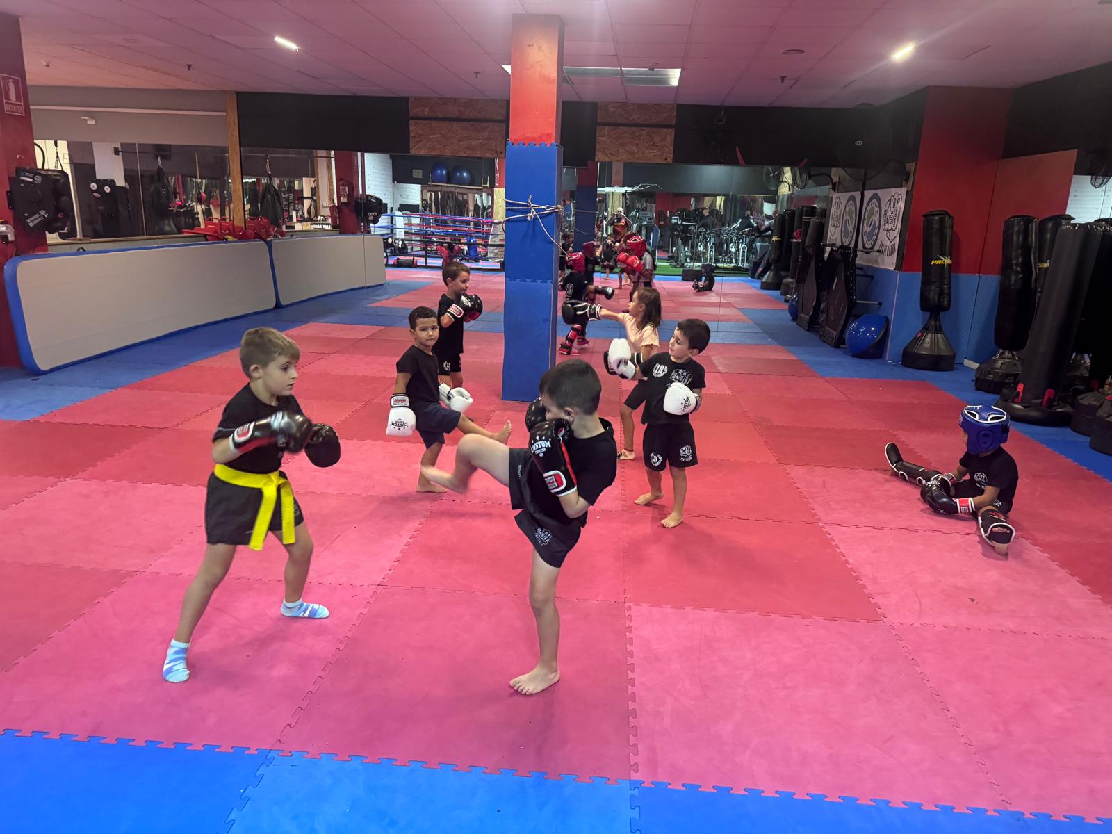 Niños y niñas practicando kickboxing con guantes y protecciones sobre el tatami de la sala de ABP Sport en Av. de Lugo, 6, con ring, sacos de boxeo y espejos al fondo